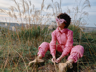 Fototapeta premium Outdoor girl in a pink outfit sits among tall grasses, wearing a playful mask, a calm reflective moment in nature amid a wide open landscape.
