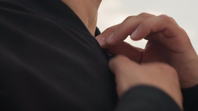 man secures coat at neck outside, male adjusting collar zipper in outdoor environment, man prepares jacket by fastening zipper near collar in outdoor setting during windy conditions