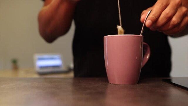 Male hands pouring sugar from a sachet into a pink mug with a hot beverage. A man stirring his morning coffee or tea in the kitchen