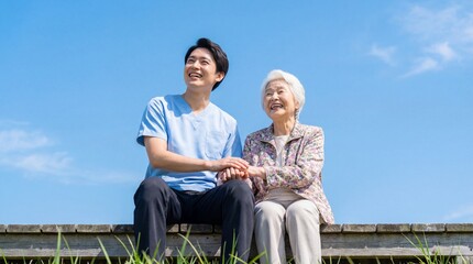 Fototapeta premium Young Asian male caregiver holding hand of senior woman sitting outdoors against blue sky