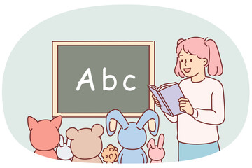 Little girl plays school teacher and reads book to toys sitting near blackboard with alphabet