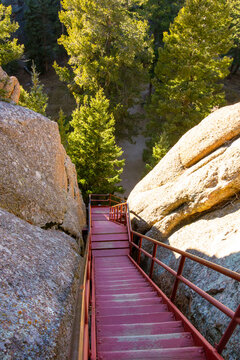 view of the iconic 143-step metal staircase leading to the historic Devil's Head Fire Lookout in Colorado. The red structure scales massive granite boulders under a bright, clear blue sky.