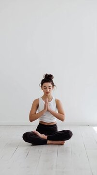Young woman meditating indoors in a bright, clean, and minimalist space. She sits in a lotus pose with hands in prayer position and eyes closed.