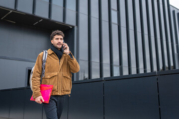 Fototapeta premium Young man walking outdoors in urban setting, talking on phone, carrying backpack and folder, modern architecture background