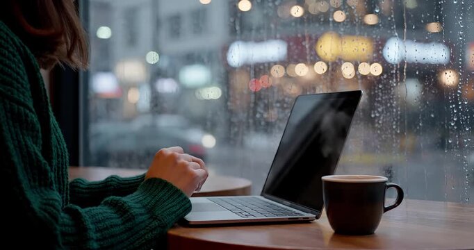 Woman typing on laptop near rainy window