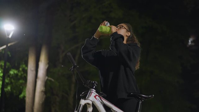 Individual Inspects Pathway Before Evening Ride, Nighttime Cyclist Examines Route Amidst Illuminated Surroundings, Female Cyclist Reviews Route Plan While Adjusting Bottle Under Bright Streetlamps