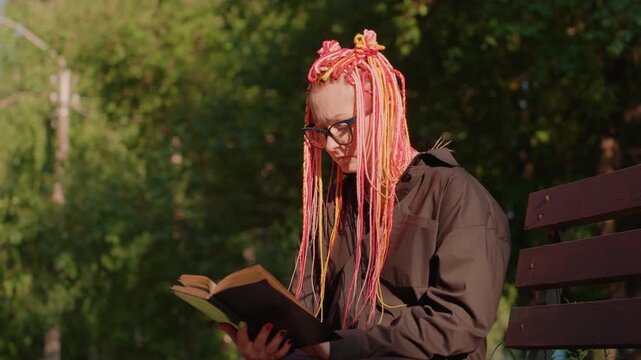 young lady engrossed in book on park bench, female reader with glasses enjoying sunny afternoon outdoors, woman with vibrant pink hair and glasses seated in sunlight reading novel peacefully