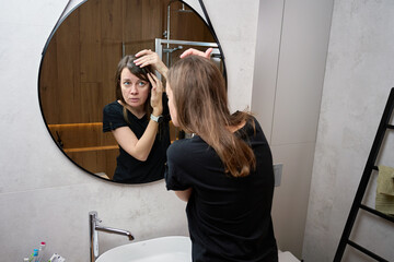 Woman looking at hairline and touching hair while standing in front of bathroom mirror.