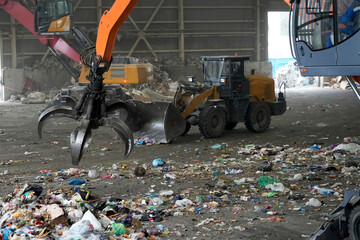 A grappling machine operates in a recycling plant, picking up and sorting through various types of household waste, including plastics and paper to aid in disposal