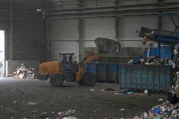 A loader uses a scoop to lift and sort household garbage and waste at a recycling plant. The indoor facility contains large bins for disposal and various debris scattered on the floor