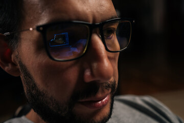 Close-up face of bearded trader male focusing on financial data and stock market trends, trading cryptocurrency and investments online, with screen reflections visible in blue light blocking glasses.