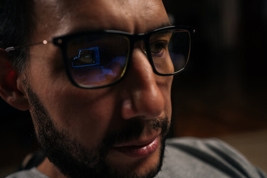 Close-up face of bearded trader male focusing on financial data and stock market trends, trading cryptocurrency and investments online, with screen reflections visible in blue light blocking glasses. - Powered by Adobe