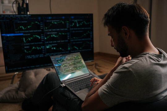 Pensive focused trader male engrossed in financial analysis and day trading, using laptop and large external monitor displaying various stock market and cryptocurrency charts from home office. - Powered by Adobe