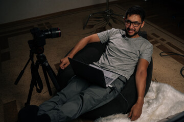 Cheerful content creator male sitting in beanbag chair, filming video blog with laptop and camera, connecting with online viewers from home workspace. Concept of modern technology.