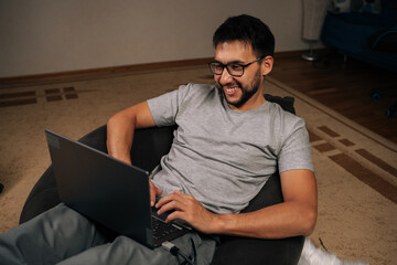 Relaxed freelancer male sitting comfortably in beanbag chair, working on laptop computer at night, smiling looking screen, enjoying convenience and flexibility of working from home.