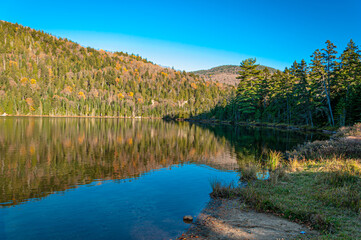 Calm lake reflecting autumn forest and mountains in Mont-Tremblant National Park, Quebec. Colorful fall foliage, clear blue sky, and peaceful wilderness landscape.
