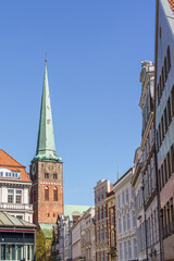 A tall green steeple with a clock on it stands in front of a row of buildings in Lubeck Germany. The sky is clear and blue, and the buildings are white