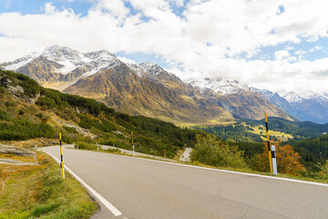 An asphalt road on the San Bernardino Pass in Switzerland The mountains are partially covered in snow. The mountains are also covered in a beautiful, colorful autumn blanket sky is blue and cloudy