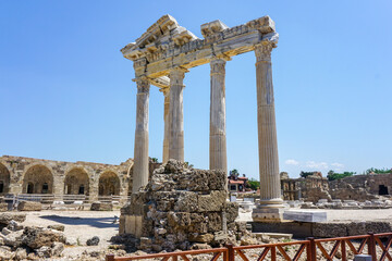 An ancient structure Apollon Temple in the form of many pillars and an arch embedded in the side of Turkey In the background is the blue Mediterranean Sea. The sky is bright and blue