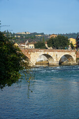 An ancient stone bridge across the Adige River in Verona, Italy. On the right side of the river. Houses are visible along the coast The sky is blue and clear.
