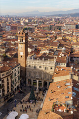 A panoramic view of the city of Verona, Italy, from above. A tall, square, red brick tower is visible in the center of the image The Alpine mountains are visible in the background