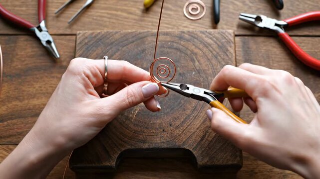 Hands work with pliers to shape wire into art. Tools spread across wooden surface. Concept of crafting, jewelry making, DIY projects