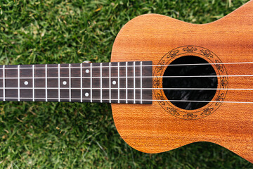 Obraz premium A classic brown acoustic ukulele with white wooden strings rests on the green grass like a folk musical instrument ready to play a jazz and rock music concert