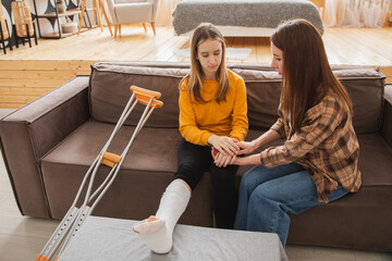 Girl with broken leg in cast sitting with mother on couch at home. Sad teenage kid recovering from broken leg. Girl using cast crutches for quick rehabilitation. Mom supporting daughter