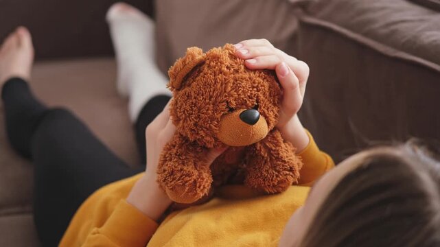 Young girl with broken leg cast sitting on sofa. Sad little kid hugging her teddy bear recovering from broken leg at home. Girl using cast and crutches. Accident injury fracture treatment foot bandage