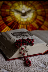 Fototapeta premium Religious still life with lit candle, open Bible, rosary. Blurred stained glass background with dove of the Holy Spirit.