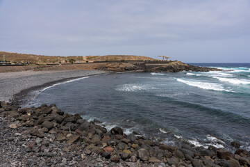Waves crash on black pebble beach at Playa del Puertito in Los Silos, with rocky breakwater, ocean bay, distant yellow structure, and cloudy horizon in Tenerife, Canary Islands, Spain.