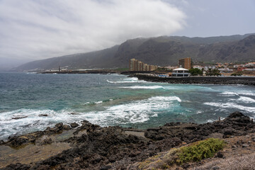 Ocean waves crash on volcanic rocky shore at Playa del Puertito in Los Silos with high-rise apartments and rugged mountains under cloudy sky in Tenerife, Canary Islands, Spain.