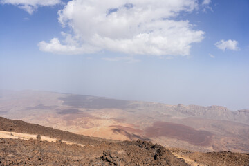 Vista from lookout point reveals Teide volcano caldera with reddish volcanic slopes, sandy expanses, rocky ridges, distant haze, scattered clouds, and blue sky in Tenerife, Canary Islands, Spain.