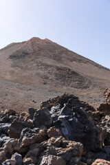 Rocky slope of Mount Teide peak rises above volcanic terrain with scattered stones and clear blue sky in Teide National Park, Tenerife, Canary Islands, Spain.