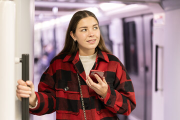 Young woman is standing in metro wagon with phone, getting to work. She is in transport, travels in underground railway transport. Girl whiles away time on trip communicating on social networks