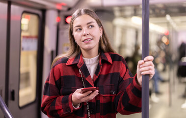In subway car, girl with phone standing in carriage, go to work, get bored on way and chatting on social networks, listen to names of stops. Public urban electric transport, daily routines of citizen
