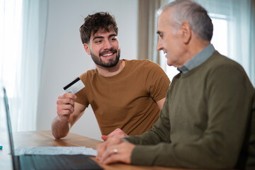 A young man assisting his senior father with online banking and card payment at home. Two generations discussing finances, bills, and digital payments in a comfortable home environment. Family support