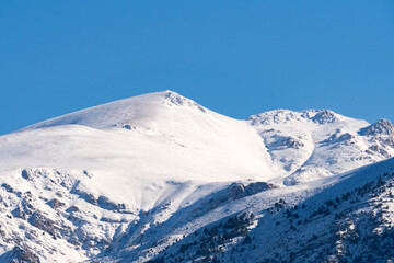 A towering mountain peak reaches into the clear blue winter sky with fresh snow covering its rocky ridges as sunlight creates high contrast textures in the frozen wilderness region area