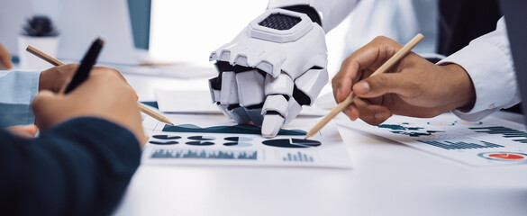 A human hand and a robot hand collaboratively analyze data reports in a modern office space,...