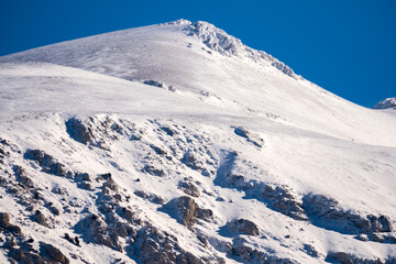 Majestic snowy mountain summit reaches into the clear blue sky under the bright winter sun featuring dramatic shadows and pristine white snow textures in a wild nature scene