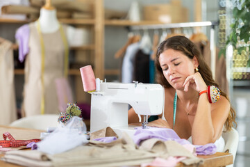 Young female dressmaker sitting at worktable in front of sewing machine with unfinished garment in...