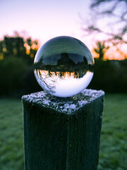 A glass ball is sitting on a wooden post in a field. The ball is reflecting the sky and the surrounding trees in a mirror-like effect. The scene is serene and peaceful