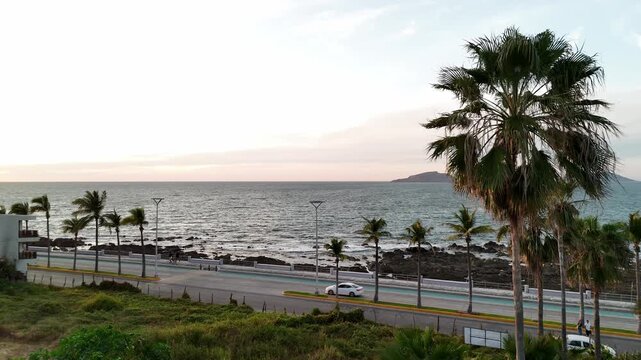 El sol en Sinaloa, iluminando el oc&eacute;ano Pac&iacute;fico y el malec&oacute;n costero. La escena muestra edificios frente al mar, palmeras movidas por el viento y el reflejo dorado del sol sobre el agua
