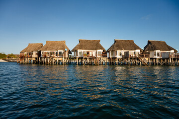A row of traditional thatched-roof bungalows built on stilts over the turquoise ocean water during a clear day in Zanzibar.