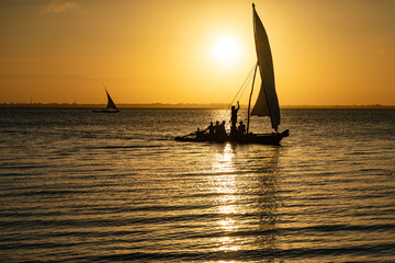Silhouette of a traditional dhow sailing boat with people on board against a golden sunset over the ocean in Zanzibar.