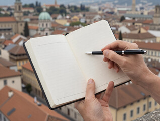 A traveler?s hand holds an open notebook and pen over a panoramic view of a historic European city.
