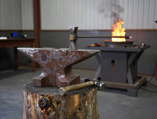 Heavy-duty anvil on a rugged tree stump with a hammer, in a metalworking shop with a glowing portable forge in the background