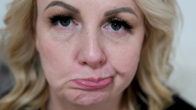Blonde woman frowns in blue studio with furrowed brow and pursed lips while facing camera with direct gaze; doubt.