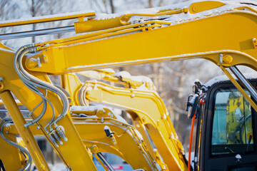 ow of bright yellow excavator arms and hydraulic lines beside cab glass, industrial construction equipment parked outdoors with snow and cold weather detail