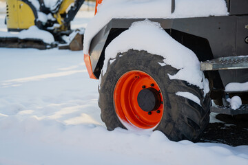 Snow covered heavy equipment tire and orange wheel rim beside metal step, close view of industrial vehicle parked outdoors in cold winter conditions